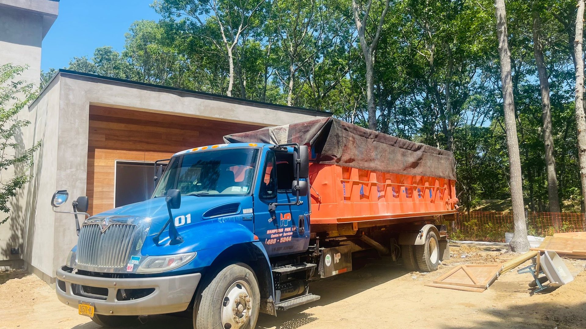 A blue Hamptons Pick Up truck delivering an orange roll-off dumpster to an East End home
