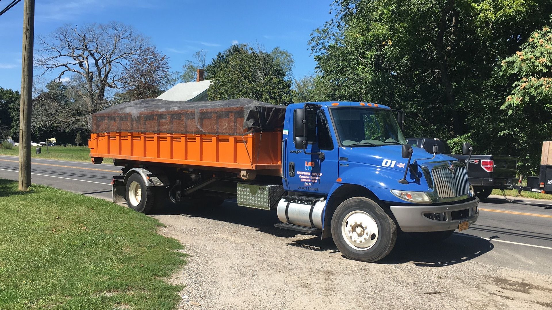 A blue Hamptons Pick Up truck carrying an orange roll-off dumpster on the East End of Long Island